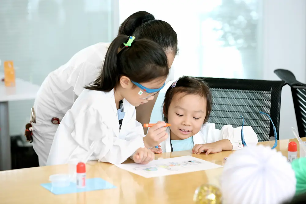 Kaksi lasta ja nainen tutkimustakissa värittämässä kuvaa pöydän ääressä