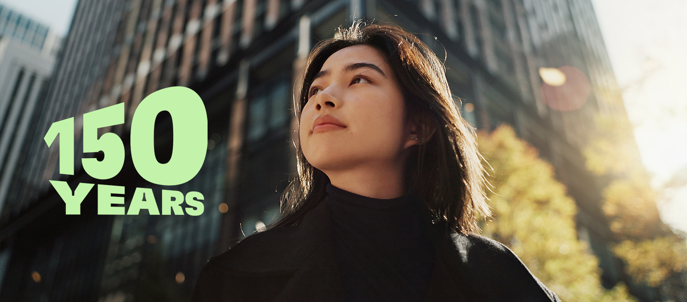 Vihreä teksti ”150 YEARS” modernin kaupunkikuvan yllä, jossa on korkeita lasirakennuksia ja syksyn puita, symboloi 150-vuotisjuhlaa.