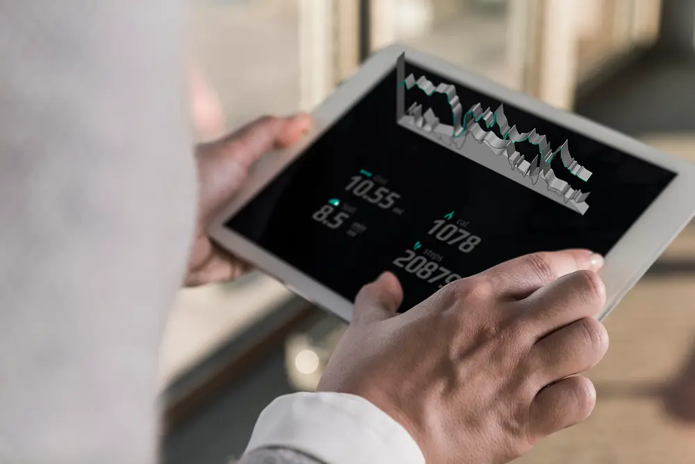 Henkilö, joka pitää kädessään tablettia, jolla näkyy digitaalisia karttoja ja numeerista tietoa; kuva on otettu olkapään yli kirkkaassa sisätilassa.
